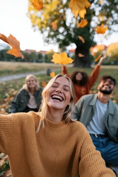 Group of friends laughing and throwing autumn leaves in a park with golden sweaters and joyful expressions during fall season
