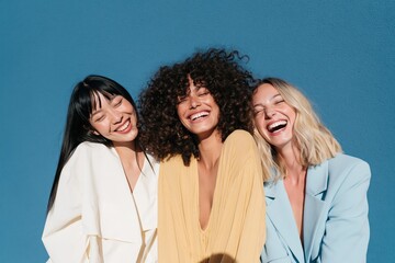 Fototapeta premium Wide group portrait of three diverse women laughing in pastel and yellow outfits against a blue textured wall for portrait layout and lifestyle projects