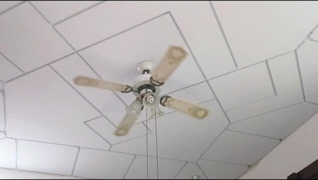 Ceiling fan with 4 blades and light fixtures spinning on a white tiled ceiling inside a residential house. Air circulation and cooling concept.