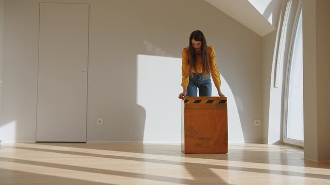 Young woman in yellow shirt and jeans carefully packing items into large cardboard box in sunlit room with wooden floor