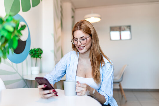businesswoman using smartphone and holding coffee cup at cafe table. Young female professional checking mobile phone messages during break in modern coffee shop. Lifestyle and technology concept