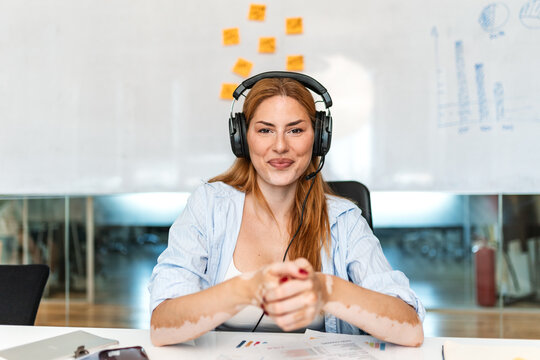 Customer service operator with headset smiling in office. Friendly female support agent sitting at desk with financial reports and whiteboard during remote assistance work. Call center concept