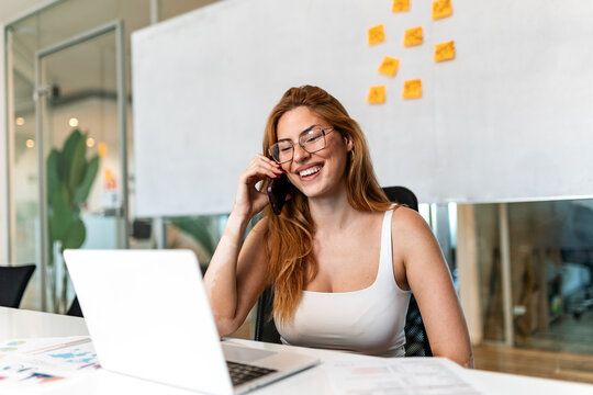 Smiling businesswoman talking on smartphone at office desk. Happy female professional making phone call while looking at laptop screen with financial documents in workplace. Communication concept