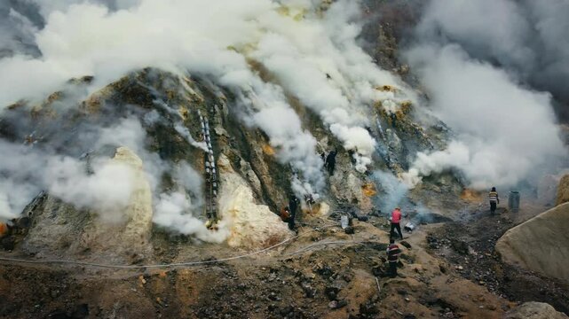 Workers navigate fumarole slopes at Kawah Ijen as white gas pours from metal pipelines and vents across the crater wall.