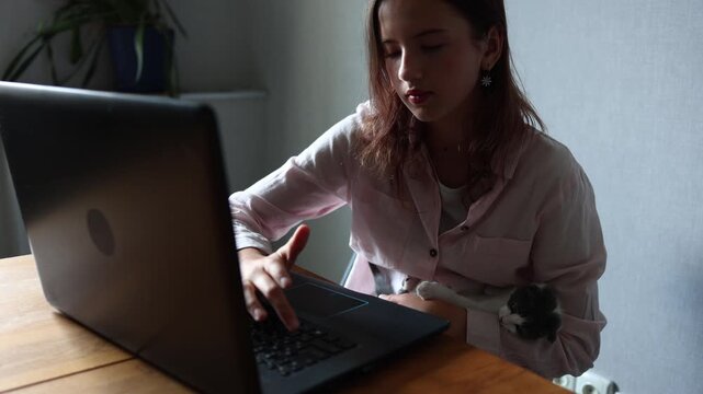 A girl sits at a wooden table with a laptop open in front of her. She holds a kitten in her arm while focusing on the screen. The room is simple, with a plant nearby.