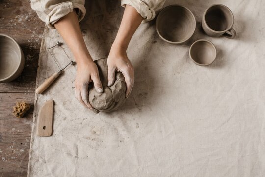 Overhead flat lay of a ceramic artist workbench with hands shaping clay, wire tool, sponge and trimmed pottery on canvas, handmade craft