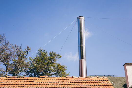 Modern metal chimney rising from a tiled roof against a clear blue sky, with green trees in the background