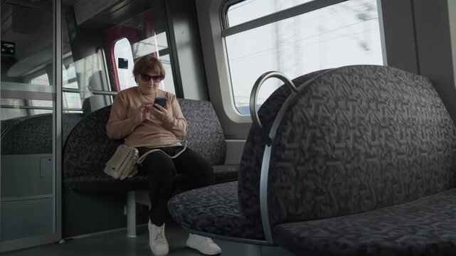 An elderly woman buys an electronic ticket on a smartphone inside a train. A mature passenger uses a mobile app for digital fare payment. Cashless online booking makes urban transit convenient.