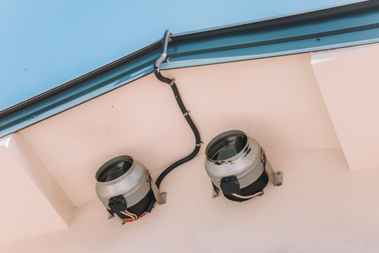 Under a building eave, two industrial inline duct fans mounted on exterior wall with conduit wiring. HVAC concept.