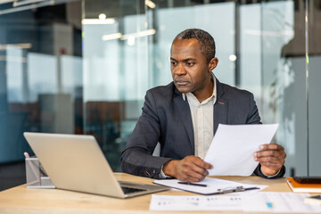 Obraz premium Experienced male executive concentrating on paperwork and financial reports at his desk, managing business tasks with a laptop in a contemporary corporate environment