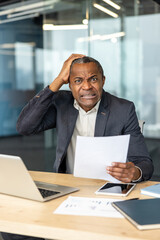 Obraz premium Frustrated adult man feeling overwhelmed and disappointed, holding a document while sitting at his desk in a modern office, grappling with a challenging situation or problem