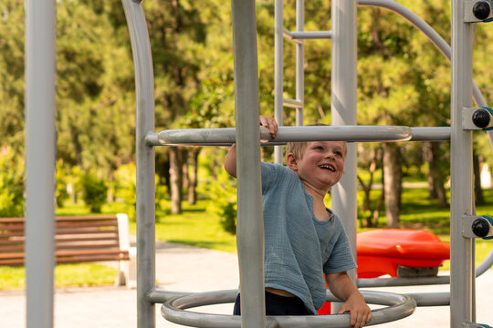 Smiling young boy holds the metal bars while moving through an outdoor playground climbing frame in a green park.