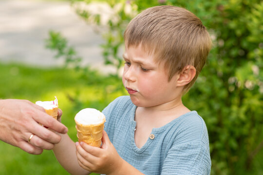 Young boy holds one vanilla ice cream cone while an adult offers another, creating a moment of choice during a summer walk in the park.