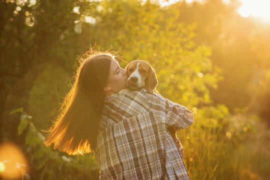 Young woman kissing her beagle while holding the dog outdoors in warm sunset light. Emotional lifestyle portrait highlighting love, trust and affectionate companionship.