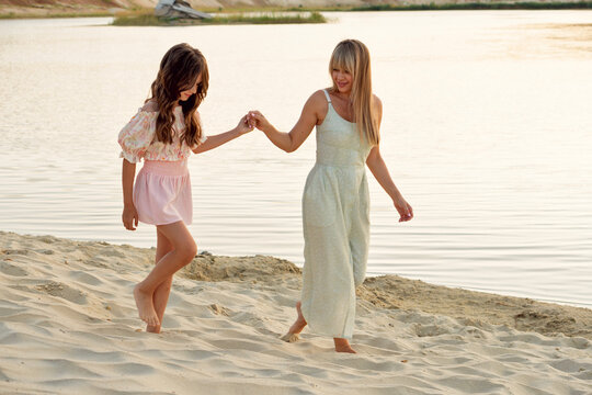 Mother and her nine-year-old daughter hold hands while walking barefoot over soft sand beside a calm lake, with pale layered cliffs and still water behind them.