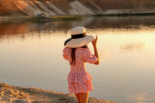 Girl stands on a sandy lakeshore adjusting a wide brim hat, viewed from behind against calm water and warm sunset reflections in a quiet outdoor setting.
