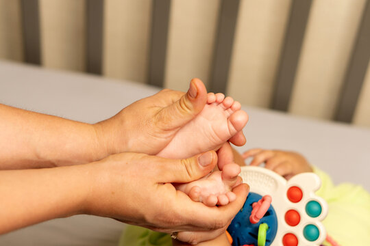 Mother gently holds and massages both bare feet of her baby son on a changing mat, with a colorful sensory toy visible in the softly focused background.