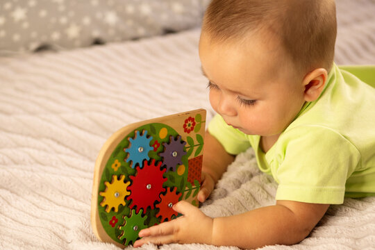 A baby boy in a lime green romper lies on his stomach and studies a colorful wooden gear puzzle on a soft bed, using both hands during sensory play and early learning at home.