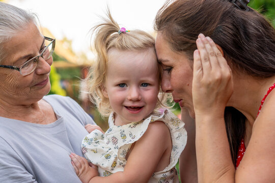 Grandmother holds her smiling young granddaughter while the mother gently touches her forehead during a tender family moment in a summer garden. Three generations