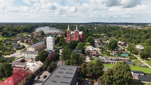 Drone video reveals the Church of St. John the Baptist in Plungė, Lithuania, surrounded by lush summer greenery, residential homes, and a scenic lake under bright daylight skies. Aerial footage.