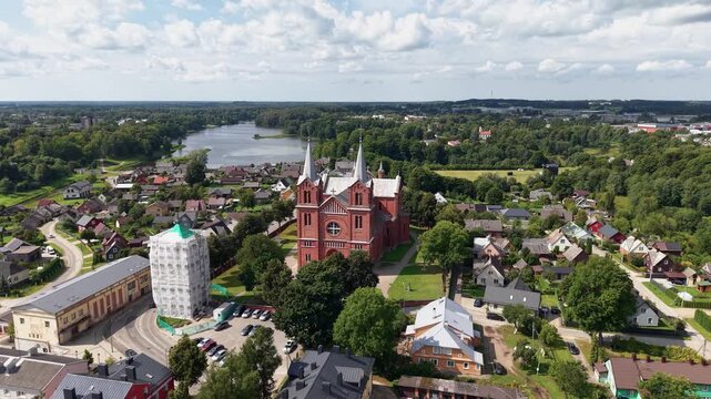 Stunning drone video showcases the Church of St. John the Baptist in Plungė, Lithuania, with red brick fa&ccedil;ade, lush greenery, village rooftops, lake and serene blue sky, captured in crisp daylight