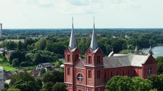 Stunning drone video captures the Church of St. John the Baptist in Plungė, Lithuania, with red brick towers, lush greenery, sparkling river, and clear daylight in a sweeping establishing shot.
