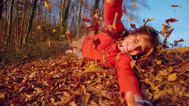CLOSE UP, PORTRAIT: Smiling woman rolls through colorful fallen leaves on forest floor while reaching toward camera. Warm autumn light, swirling foliage and moment filled with laughter and happiness.