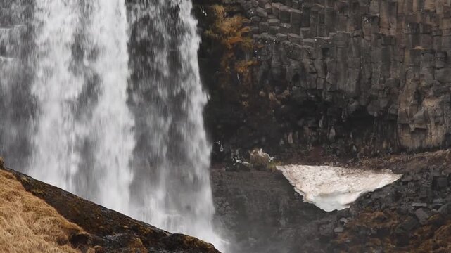 Static close-up of Svodufoss waterfall plunging past dark basalt columns beside snow patch, Rif, Snaefellsnes