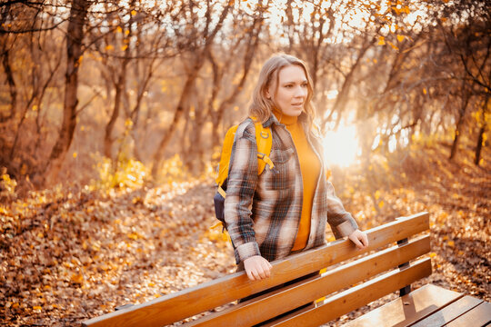 Woman hiker resting near bench in golden autumn park &mdash; outdoor adventure and solo travel concept. Active lifestyle, autumn season wellness. Relaxed female tourist enjoying nature walk.