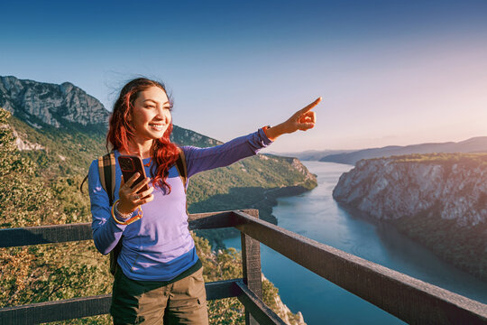 Smiling female traveler with smartphone points toward a winding Danube river Iron Gates gorge from a wooden overlook, discovering her next destination. Wanderlust concept.