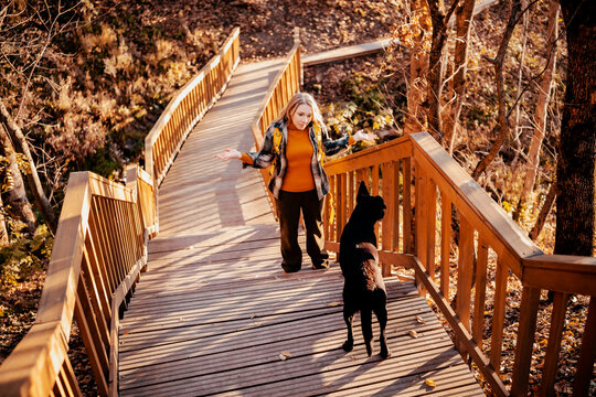 Young woman with dog on wooden park bridge &mdash; pet owner behavior and training concept. Puzzled female hiker shrugging hands during autumn walk. Active outdoor lifestyle with domestic animal.