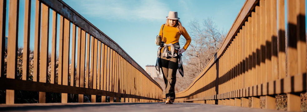 Active woman on autumn hike walking across wooden boardwalk - outdoor adventure. Female traveler exploring nature park with coffee cup. Physical well-being and active lifestyle trend.