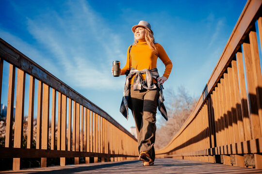Smiling woman in autumn park strolling on wooden path with hot drink - weekend getaway. Cheerful female hiker enjoying sunny outdoor weather. Active recreation and solo travel trend.