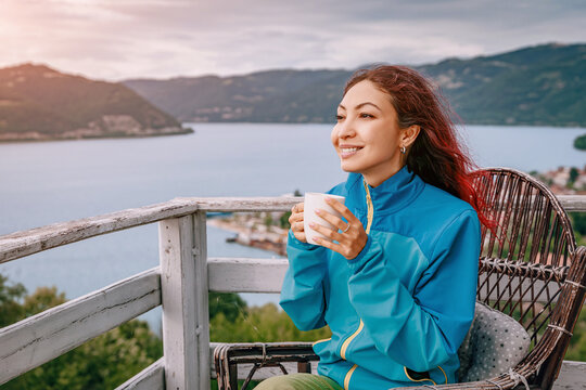 Rustic terrace railing and panoramic lake basin frame a smiling woman sipping coffee outdoors. Relaxation concept.