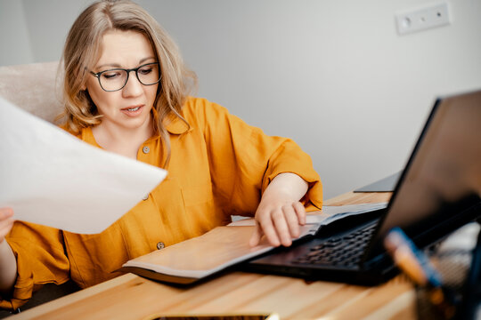 Focused woman in glasses reading documents near laptop &mdash; home office work and financial audit concept. Concentrated female employee analyzing reports at workplace, remote job stress, paperwork.