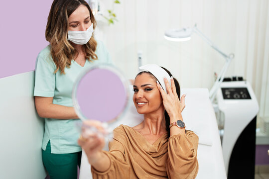 Smiling female client admiring her face reflection in a handheld mirror while visiting a professional aesthetician in a medical spa or clinic for rejuvenation procedures.