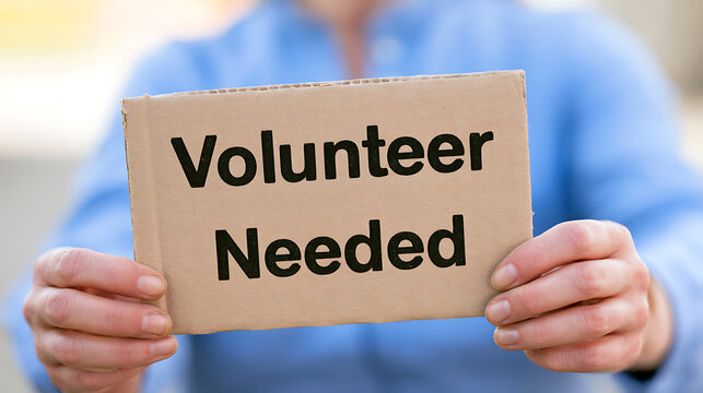 A close up view of a person holding a cardboard sign with the text volunteer needed written on it in black ink for community outreach