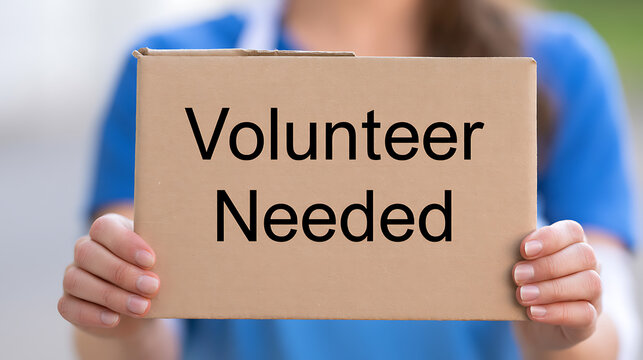 A close up view of a person holding a cardboard sign with the text volunteer needed written on it, representing community service and aid