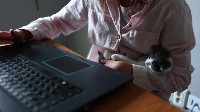 A girl sits at a wooden table with a laptop open in front of her. She holds a kitten in her arm while focusing on the screen. The room is simple, with a plant nearby.