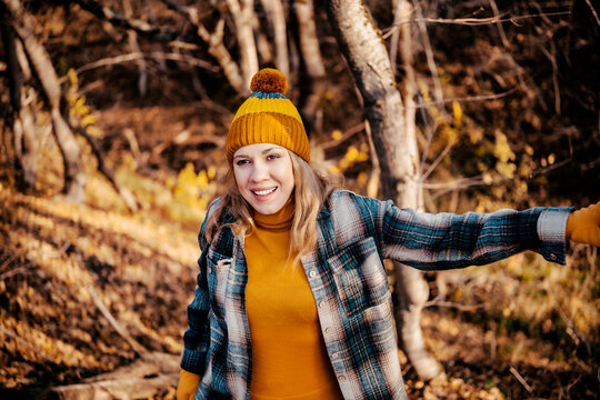 Joyful woman in knitted beanie hiking in autumn forest &mdash; outdoor wellness and fall season travel concept. Smiling female tourist enjoying nature walk, active lifestyle and positive mental health.