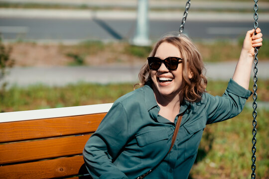 Cheerful woman in sunglasses sitting on outdoor swing &mdash; inner child and carefree summer lifestyle concept. Laughing female tourist swinging in city park, positive energy and mental well-being.