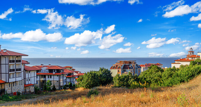 A coastal resort village features red-roofed buildings set against a calm blue sea under a bright, cloud-dotted sky.