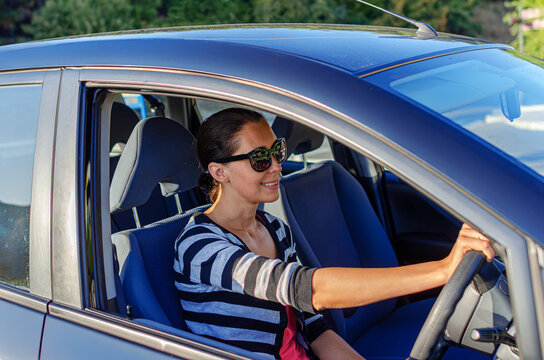 A smiling woman wearing sunglasses holds the steering wheel while driving a car on a sunny day.