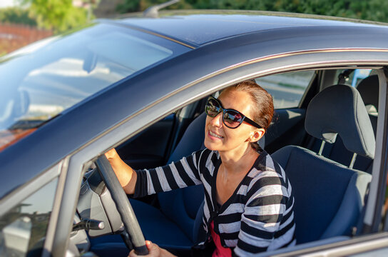 A smiling woman wearing sunglasses holds the steering wheel while driving her car on a sunny day.
