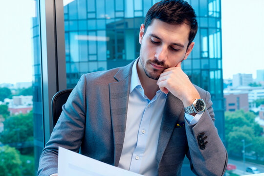 Young adult Caucasian businessman sitting in office, reading documents with focused expression. Image represents corporate decision making, suitable for financial services marketing
