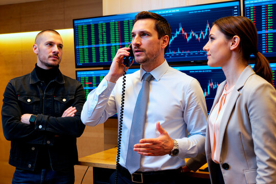 Diverse group of young adult financial analysts tracking stock market data on screens while colleague talks on phone. Image suitable for corporate trading and investment marketing