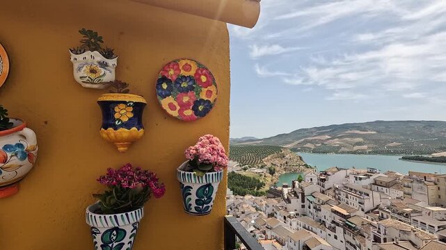 Painted ceramic wall pots on an ochre wall, pan revealing Iznajar and its reservoir from Mirador de San Rafael, Cordoba, Spain