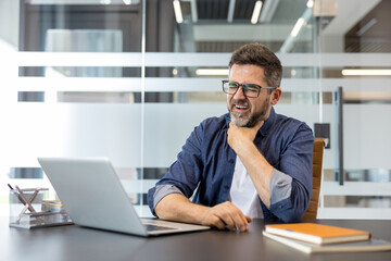 Obraz premium Man with glasses at office desk rubbing his throat in discomfort, looking sick and exhausted while trying to work on a laptop during illness or flu symptoms, stressed and unwell