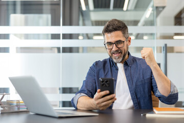 Fototapeta premium Happy businessman at his office desk clenches his fist in excitement while reading good news on his smartphone, celebrating a win and smiling at the notification on screen