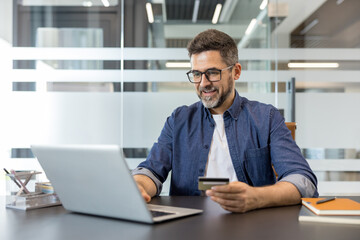 Obraz premium Man holding a credit card while using a laptop for secure online shopping and e-commerce transactions from a modern office desk, confident and smiling during remote work